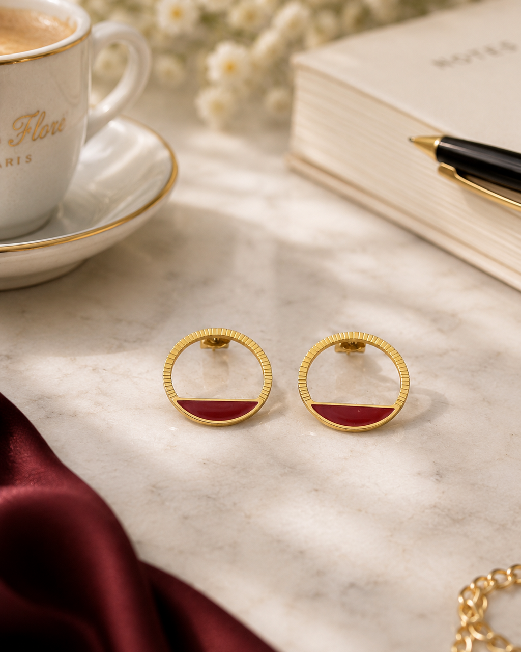détails des boucles d'oreilles Livia dorées en acier inoxydable avec une touche de rouge profond.Les boucles d'oreilles sont posées sur une table