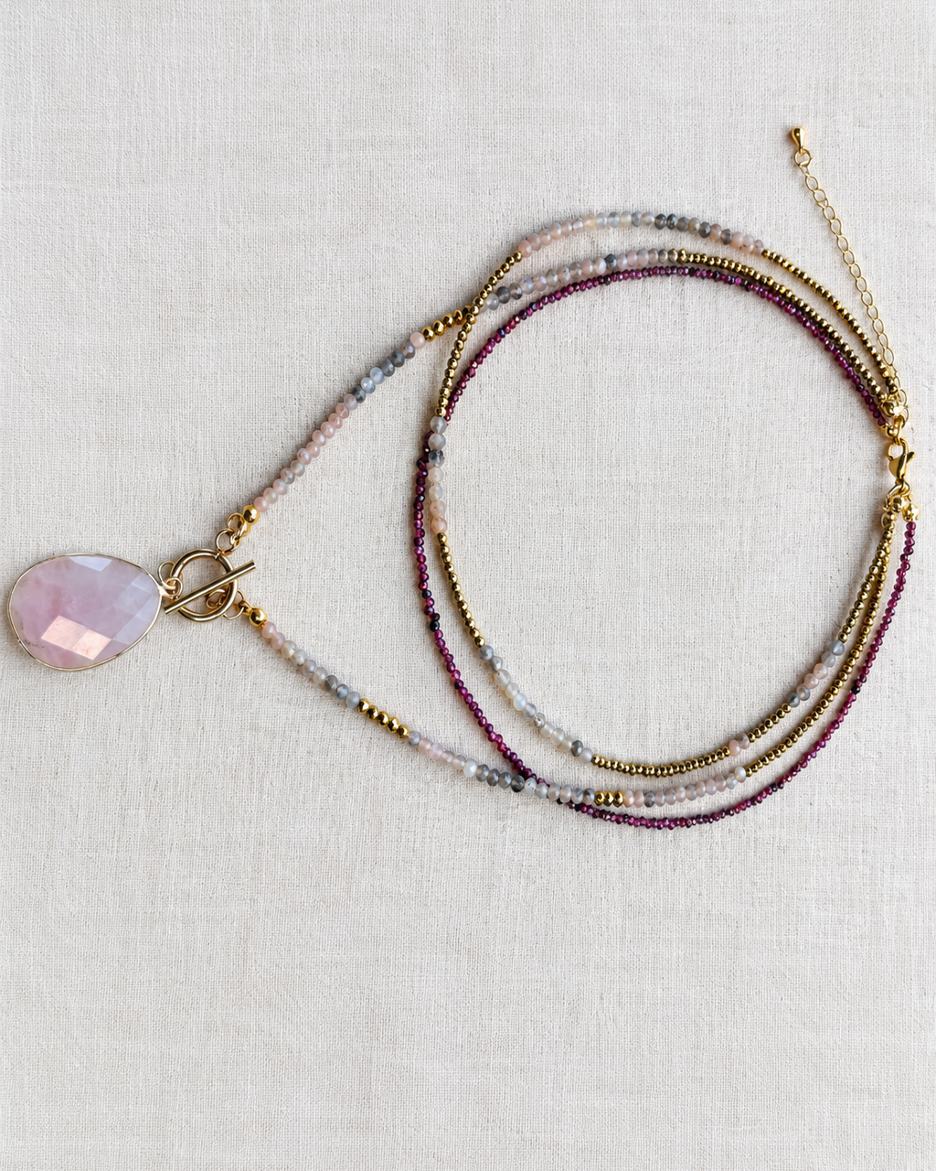 Collier trois rangs Juste avant l'aube posé sur un tissu blanc, associant quartz rose, rubis, pierre de soleil noire et hématites dorées. Créé et assemblé en France.