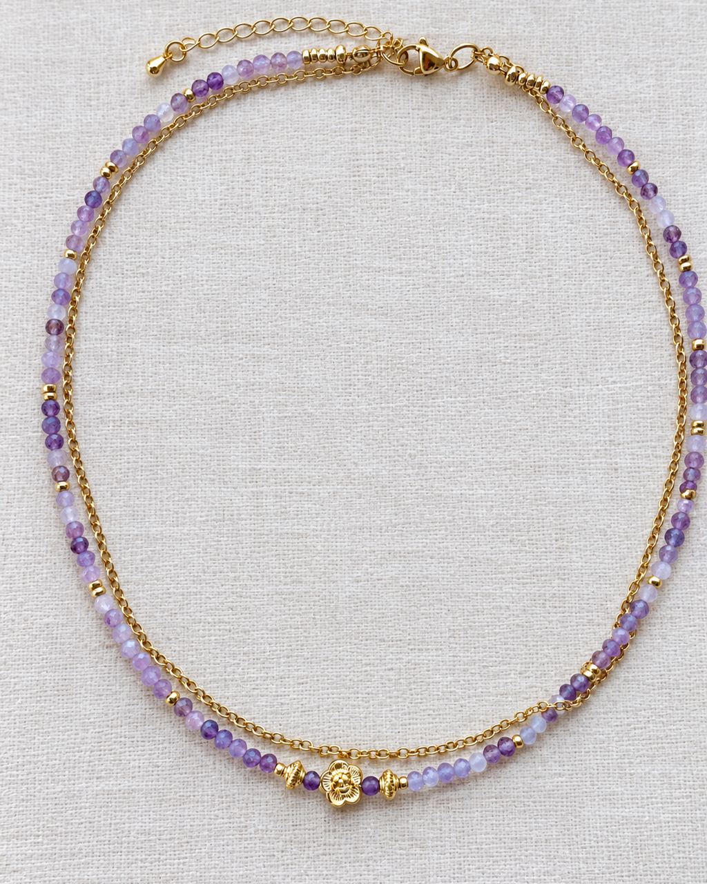 Collier double en pierres naturelles d’améthyste aux nuances violettes et lilas, associé à une chaîne plaqué or 18 k et une fleur centrale dorée, présenté sur un tissu en lin écru.ée 