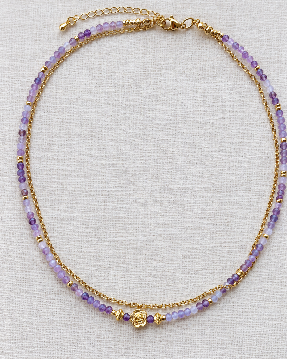 Collier double en pierres naturelles d’améthyste aux nuances violettes et lilas, associé à une chaîne plaqué or 18 k et une fleur centrale dorée, présenté sur un tissu en lin écru.ée 