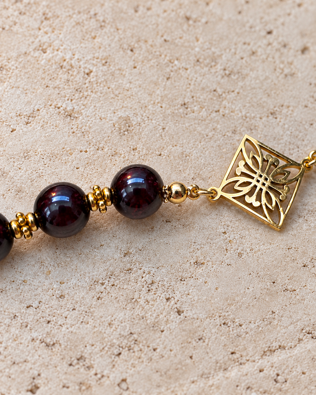 Gros plan sur un collier artisanal en perles de grenat naturel rouge profond, ornées d'intercalaires dorés et d'un pendentif carré ajouré à motif floral. Bijou créé et assemblé à la main en France dans l'atelier Yselia.