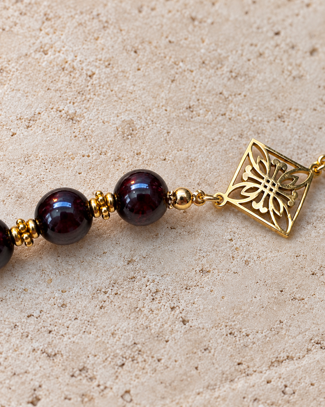 Gros plan sur un collier artisanal en perles de grenat naturel rouge profond, ornées d'intercalaires dorés et d'un pendentif carré ajouré à motif floral. Bijou créé et assemblé à la main en France dans l'atelier Yselia.
