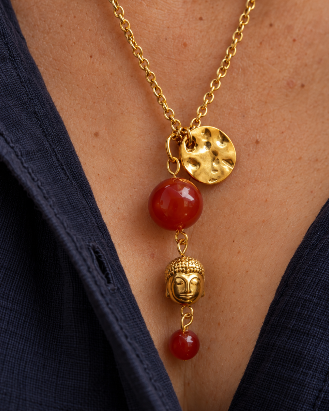 Gros plan sur le pendentif du sautoir Namasté, composé d'un Bouddha en hématite dorée, de deux perles de cornaline rouge orangé et d'un médaillon martelé en plaqué or 18 carats.