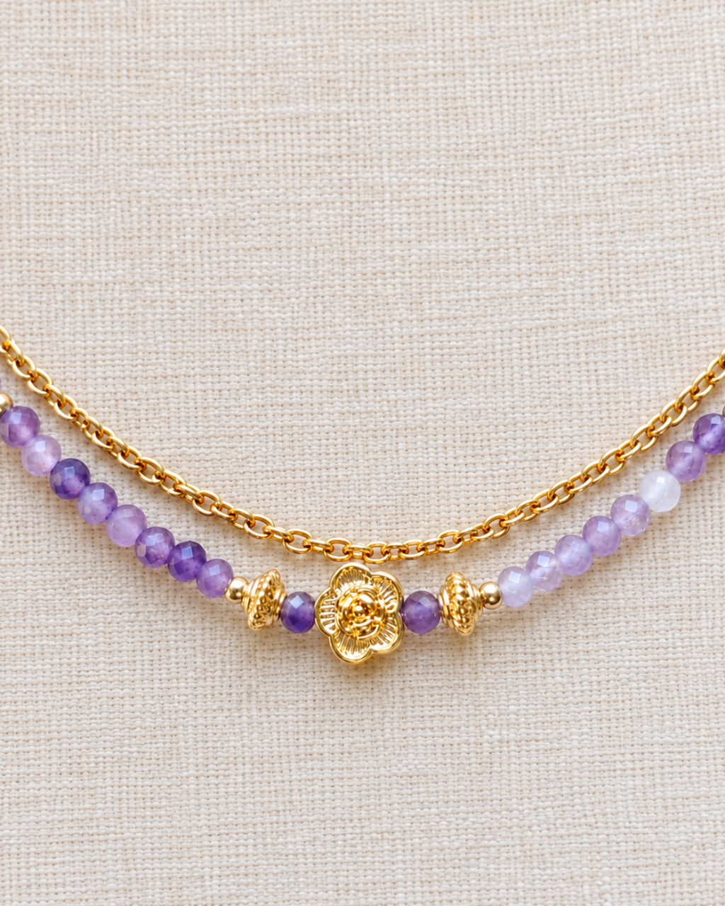 Gros plan sur le pendentif fleur en plaqué or 18 carats d’un collier en améthyste naturelle, entouré de perles violettes facettées et de détails dorés, sur un tissu en lin écru.