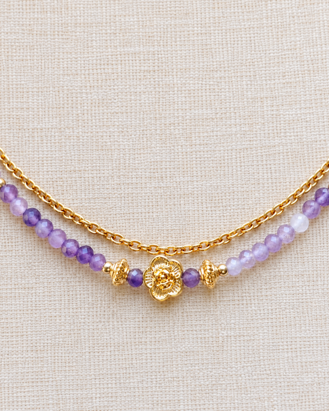 Gros plan sur le pendentif fleur en plaqué or 18 carats d’un collier en améthyste naturelle, entouré de perles violettes facettées et de détails dorés, sur un tissu en lin écru.