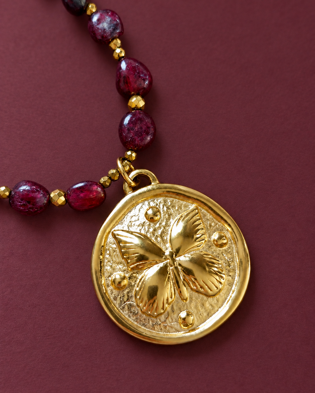 Gros plan sur le médaillon travaillé en plaqué or 18 carats du collier Les Heures Pourpres, accompagné de perles de grenat facettées et d'hématites dorées.