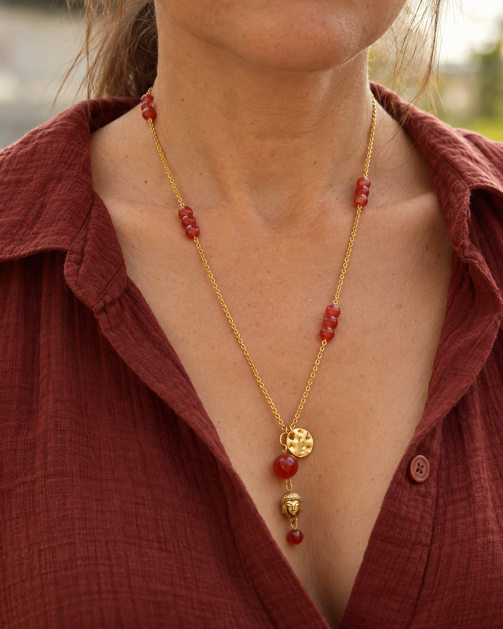Sautoir Namasté porté sur une femme, composé d'une chaîne en plaqué or 18 carats, d'une cornaline rouge orangé, d'un pendentif Bouddha en hématite dorée et d'un médaillon martelé doré.