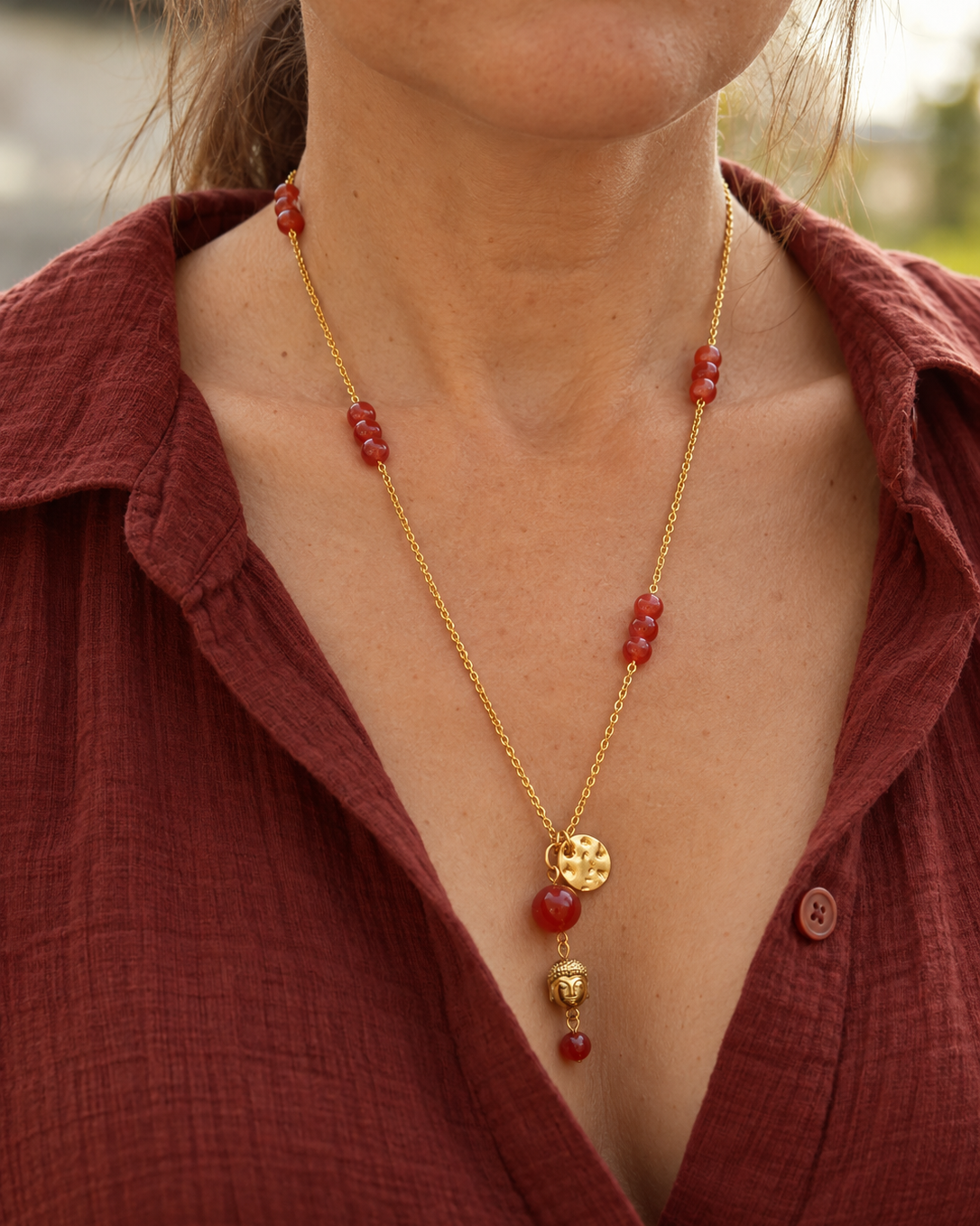 Sautoir Namasté porté sur une femme, composé d'une chaîne en plaqué or 18 carats, d'une cornaline rouge orangé, d'un pendentif Bouddha en hématite dorée et d'un médaillon martelé doré.